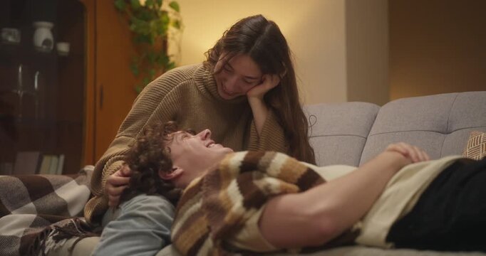 Happy girl looking at her boyfriend with curly hair who is lying on her lap and looking at her while relaxing on a cozy sofa at home in the evening