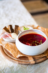 Traditional borscht soup with herbs served in a bowl on a rustic wooden tray, accompanied by rye bread, garlic, bacon, and sour cream. Eastern European homemade comfort food.