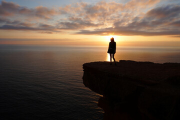 A solitary figure watches the sunset from a cliff overlooking the ocean.