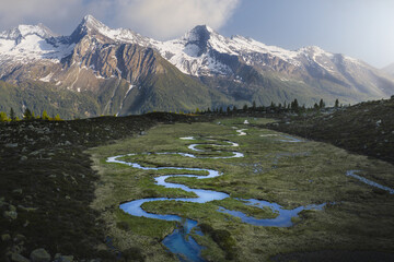 Aerial view of a serpentine river winding through a verdant valley floor towards snow-capped peaks of the Dolomites, Prettau, Trentino-South Tyrol, Italy.