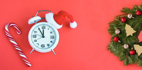 White alarm clock with Santa hat and Christmas wreath on a red background. Copy space. Close-up.