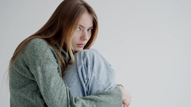 A thoughtful young woman wraps herself in a soft, thick sweater, conveying introspection and comfort against a plain white background. Attractive woman with long hair posing for camera.