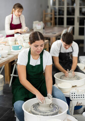 Interested young woman in pinafore making pottery product by potter wheel in art studio. Persons...