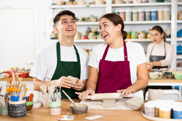 Cheerful young man and woman in aprons modeling handmade mugs of row clay material in pottery workroom