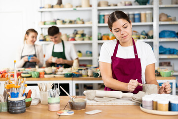 Fototapeta premium Positive young woman in pinafore cutting the edge of row clay cup with filament in art studio