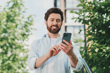 Young man in casual shirt using smartphone outdoors in urban setting surrounded by greenery and...