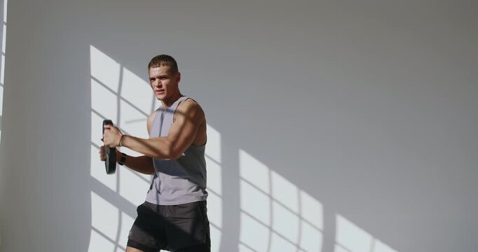 A 23-year-old man with an athletic build is engaged in weight training in a well-lit studio. Young Athletic Man Lifting Weights in a Sunlit Studio Setting