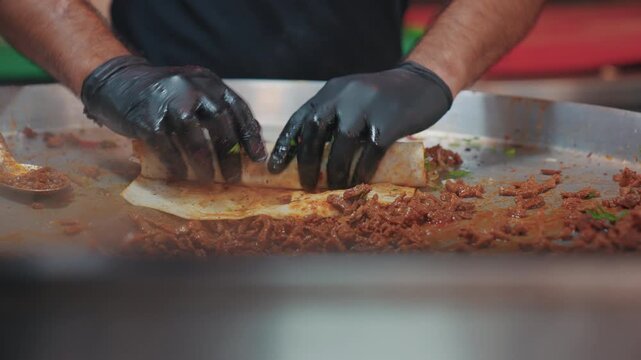 Close-up of a chef with black gloves preparing a traditional Turkish tantuni wrap. The cook is rolling spicy minced meat in flatbread (lavaş) on a hot griddle (sac).