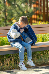 Little stylish schoolboy in blue bomber jacket sits on a bench with her backpack after school...