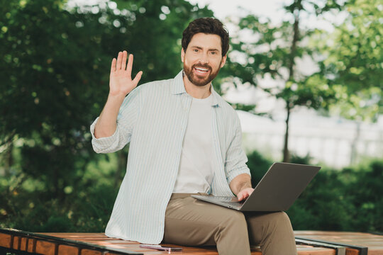 Young man outdoors using a laptop and waving with a friendly smile, reflecting a casual and productive lifestyle. - Powered by Adobe