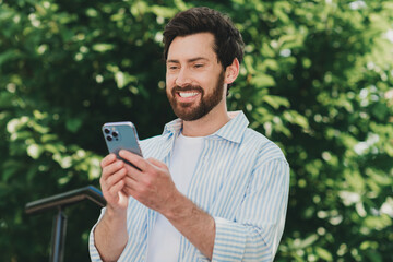 Handsome man smiling while using a smartphone outdoors in sunny weather surrounded by greenery