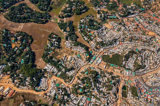 Aerial view of an expansive settlement with tightly packed structures contrasting with the surrounding green fields and winding paths, Balukhali Bazar, Chittagong Division, Bangladesh.