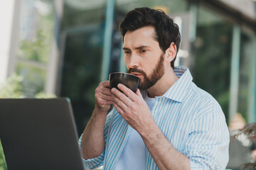 Attractive man enjoys coffee outdoors while working on a laptop, embodying casual city lifestyle...