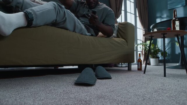 Ground level shot of bored young Black man under house arrest lying in front of TV with empty beer bottles around, getting off couch, putting on slippers and going away