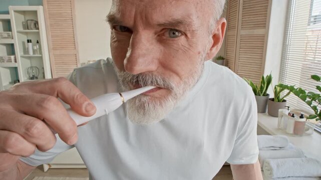 Tilt up of aged bearded man wearing white t-shirt squeezing toothpaste on electric toothbrush over porcelain sink and starting brushing his teeth in front of mirror in bathroom