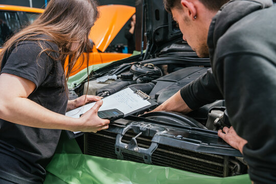 Young male and female mechanics collaborating on car engine inspection with a form and mobile phone
