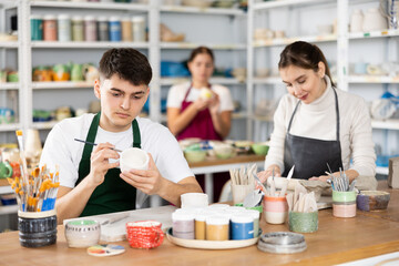 Concentrated young man in apron painting on handmade clay cup with brush in pottery workshop....