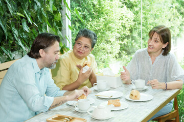 Asian senior woman and two Caucasian friends sit at a cafe table, enjoying pastries and tea while...