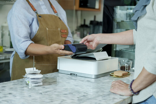 Asian senior female barista adult holding terminal while customer taps credit card for cashless payment at cafe counter reflecting efficient service in local small coffee shop
