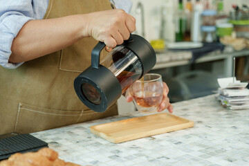 Asian senior female barista adult pouring freshly brewed tea from modern teapot into glass at cafe...