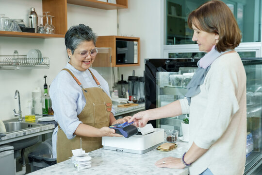 Asian senior female barista adult assisting customer tapping credit card on payment terminal at cafe counter showing efficient cashless payment process in local family coffee shop