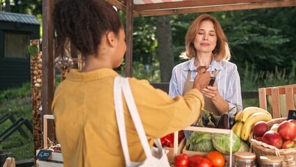 Rear of young African American woman buying food at market outdoors paying with credit card. Caucasian female farmer selling organic farm vegetables at food market counter. Buyer concept