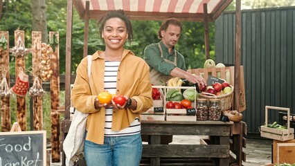 African American woman outdoor buying food at food market. Woman buy vegetables and paying with smartphone pay pass. Male and female family of farmers work at market selling organic products