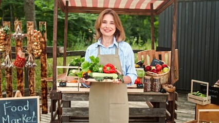Camera approaching mature beautiful joyful woman in apron standing outdoor holding fresh vegetables and smiling to camera. Caucasian happy female farmer with food at selling local market