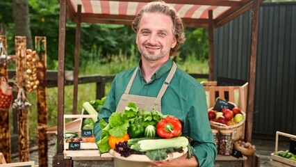 Close up of handsome Caucasian mature man farmer in apron standing outdoor holding in hands board with fresh green vegetables looking and smiling to camera. Male selling organic vegetables at market
