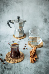 Black coffee in curved glass with water tumbler, moka pot and cinnamon sticks on woven coasters, scattered beans on pale stone background, airy vertical layout for cafe ads