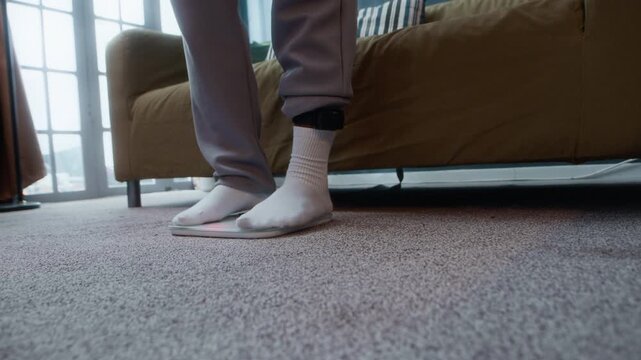 Cropped ground level shot of unrecognizable man under house arrest wearing electronic monitoring ankle bracelet weighing himself stepping on scales on floor