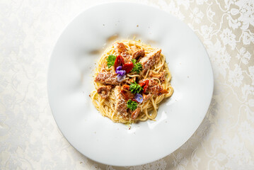 Spaghetti carbonara garnished with cherry tomatoes, parsley, and edible flowers on a white plate with grated cheese and meat, served on an elegant tablecloth.