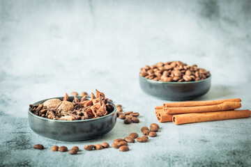 Bowls with aromatic winter spices and roasted coffee beans, cinnamon sticks and scattered beans on pale stone, airy layout for seasonal beverage or recipe designs