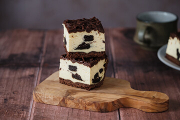 Slices of rich cheesecake with layers of chocolate and cookies stand on a rustic wooden board. The warm, inviting atmosphere enhances the dessert's appeal.