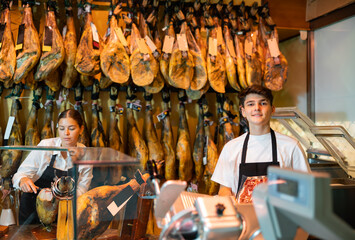 Skilled young salesgirl and salesman of butcher shop selling delectable dried Iberian jamon, working together at counter