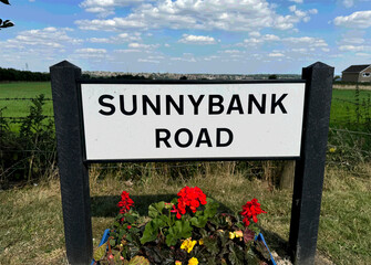 Sunnybank Road sign framed by summer flowers and soft clouds in Mirfield, West Yorkshire, UK.
