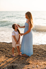 Smiling woman and kid daughter holding hands walking at beach over sea outdoors. Summer vacation season. Motherhood.