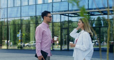 Colleagues are talking while standing outdoors near an office building. They drink coffee from...