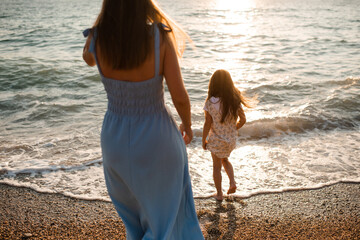 Mother and child daughter holding hands walking at beach over sea outdoors. Summer vacation season. Motherhood.