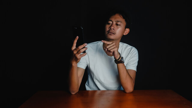 A young man wearing a white t-shirt sitting at a table, intensely staring at his smartphone in a dark studio setting., Concept of focus, concentration, social media, or technology addiction.