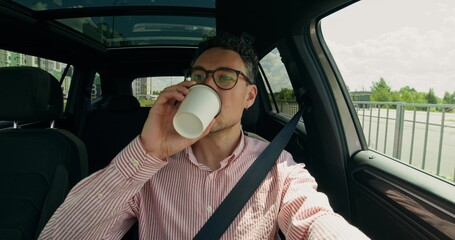 Front view of a stylish man driving a car. He drinks coffee from a disposable cup.
