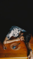 A young man looking exhausted or stressed, resting his head on a wooden table with an empty plate...