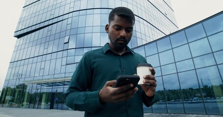 A man uses a mobile phone and drinks a takeaway coffee while walking along a city street. Wide...