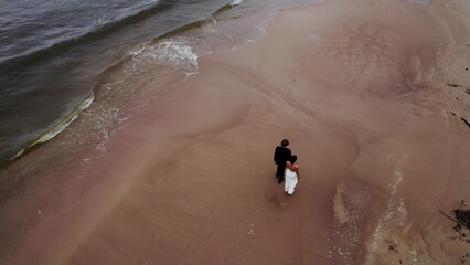 The newlyweds are walking on the beach. Sea waves are beating against the shore. View from the drone