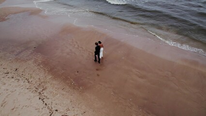 The newlyweds are walking on the beach. Sea waves are beating against the shore. View from the drone