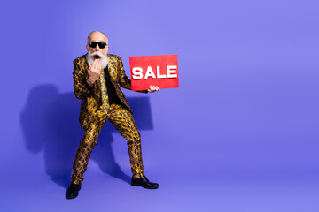 Eccentric elderly man holding sale sign in stylish outfit on bright purple background showcasing a...