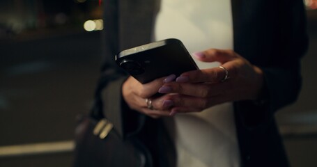A woman uses a mobile phone while standing on a city street in the evening. Close-up of her hands, unrecognizable person