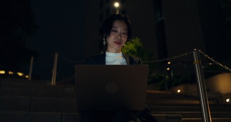 A happy woman uses a laptop and drinks coffee from a disposable cup while sitting outside at night