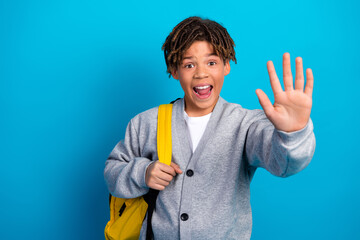 Excited young schoolboy with a backpack making a friendly hand gesture against a bright blue...