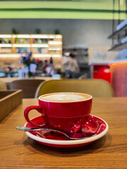 A vibrant red coffee cup filled with cappuccino sits on a wooden table. Cozy cafe ambiance in the blurred background with warm lighting creating a relaxed atmosphere.